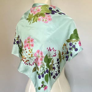 May include: A square scarf in a light mint green color featuring a botanical print of pink flowers, green leaves, and dark purple blackberries. The design is arranged in a floral pattern across the fabric, which is draped over a mannequin form.