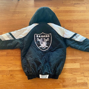 Puede incluir: Chaqueta negra con capucha y el logo de los Raiders en la espalda. La chaqueta tiene detalles blancos y grises en las mangas y un dobladillo negro. El logo de los Raiders es un escudo con un pirata con parche en el ojo.