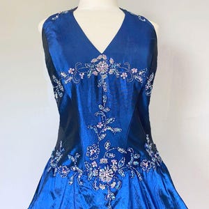 Op de afbeelding: Een mouwloze, koningsblauwe avondjurk met een V-hals. Het lijfje en de rok zijn versierd met bloemenborduurwerk in roze, zilver en blauw. De jurk wordt getoond op een witte mannequin.