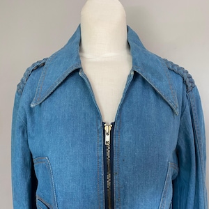 May include: A light blue denim jacket with a zipper closure and a wide collar. The jacket features braided shoulder accents and visible stitching. The jacket is displayed on a mannequin.