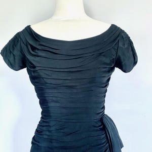 May include: A black, vintage-style dress with a ruched bodice and short sleeves. The dress features a boat neckline and a gathered waist, creating a flattering silhouette. The fabric appears to be a soft, flowing material.