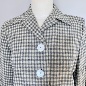 May include: A blazer featuring a grey and off-white gingham check pattern. The garment has a notched lapel collar and two large, round, white buttons centered on the front closure. The fabric has a textured, woven appearance throughout the piece.