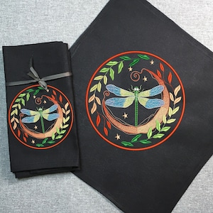 Op de afbeelding: Set van zwarte stoffen servetten met geborduurde libellenmotieven. De libel bevindt zich op een halve maan met groene bladeren en sterren. De servetten zijn met een zwart lint aan elkaar gebonden.