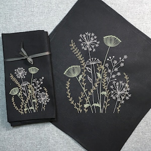 Puede incluir: Dos toallas de cocina negras con un diseño bordado de flores silvestres blancas y verdes. La toalla más grande tiene la palabra "Pteris" impresa en blanco sobre un círculo negro en la esquina inferior derecha.