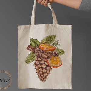 Może przedstawiać: Biała płócienna torba na zakupy z haftowanym wzorem szyszki, cynamonowych patyczków, anyżu gwiazdkowego i plastrów pomarańczy. Wzór jest w odcieniach brązu, zieleni, pomarańczy i żółci.