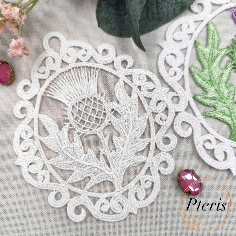 Puede incluir: Tapete de encaje blanco con un dise&ntilde;o detallado de cardo. El intrincado patr&oacute;n floral est&aacute; rodeado por un borde decorativo. La imagen tambi&eacute;n incluye una hoja verde parcialmente visible y acentos florales rosas, con la palabra "Pteris" en un c&iacute;rculo.