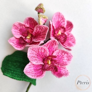 Puede incluir: Primer plano de una flor de orqu&iacute;dea rosa con una hoja verde. La orqu&iacute;dea est&aacute; bordada con puntadas detalladas y tiene un centro dorado.