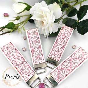 May include: Four white key fobs with red embroidered floral patterns. Each fob has a different design, including roses and geometric shapes. Silver-colored hardware secures the fob. The word "Pteris" is in a gold circle in the lower left corner.