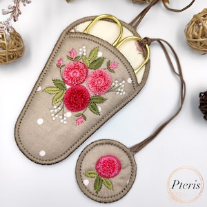 Puede incluir: Un porta tijeras de tela beige con un diseño de bordado floral con flores rosas y hojas verdes. El porta tijeras tiene un patrón de lunares blancos y un cordón marrón. Un pequeño círculo redondo de tela beige con un diseño de bordado de flor rosa está unido al cordón.