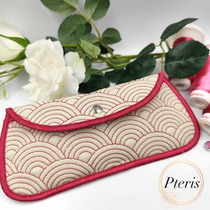 Puede incluir: Estuche para gafas color crema con un patrón de ondas rojas y ribete rojo. El estuche tiene un cierre de solapa con un broche plateado. Decorado con una rosa blanca y carretes de hilo rosa.