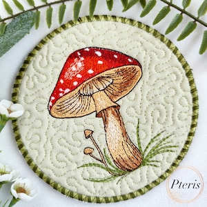 以下が含まれることがあります： 中央に赤と白のキノコが刺繍された、丸い緑色の布製のコースター。キノコは茶色の茎と赤い傘に白い斑点があります。