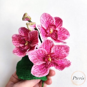 Puede incluir: Un ramo de orqu&iacute;deas bordadas de color rosa con detalles dorados en una maceta de fieltro verde.