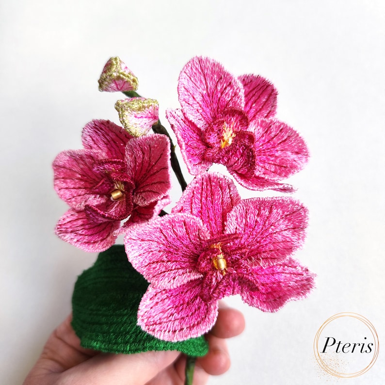 Puede incluir: Un ramo de orqu&iacute;deas bordadas de color rosa con detalles dorados en una maceta de fieltro verde.