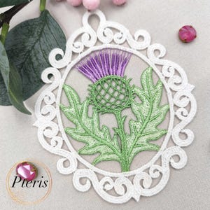 Puede incluir: Adorno de cardo bordado con una flor morada, hojas y tallo verdes, encerrado en un marco circular blanco y ornamentado. El dise&ntilde;o se encuentra sobre un fondo neutro, con la palabra "Pteris" en la esquina inferior izquierda.