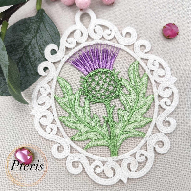 Puede incluir: Adorno de cardo bordado con una flor morada, hojas y tallo verdes, encerrado en un marco circular blanco y ornamentado. El dise&ntilde;o se encuentra sobre un fondo neutro, con la palabra "Pteris" en la esquina inferior izquierda.