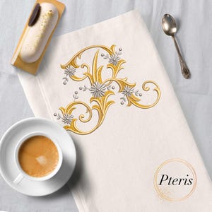 May include: Ivory-colored linen towel with a gold embroidered letter "A" design, accented with silver floral details. A cup of coffee, a pastry, and a spoon are arranged around the towel. The towel has the word "Pteris" in a gold circle.
