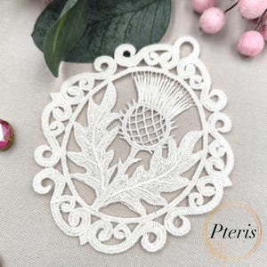 Puede incluir: Un posavasos de encaje blanco con un dise&ntilde;o detallado de cardo dentro de un marco circular decorativo. El posavasos tiene un delicado patr&oacute;n calado. La palabra "Pteris" es visible en la esquina inferior derecha.