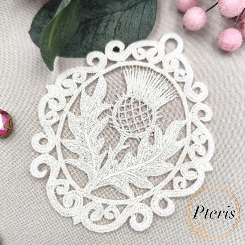 Puede incluir: Un posavasos de encaje blanco con un dise&ntilde;o detallado de cardo dentro de un marco circular decorativo. El posavasos tiene un delicado patr&oacute;n calado. La palabra "Pteris" es visible en la esquina inferior derecha.
