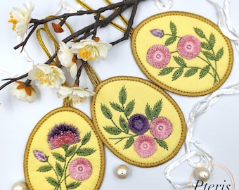 May include: Three oval-shaped yellow fabric ornaments featuring embroidered floral designs in pink, purple, and green. Each ornament has a gold-colored stitched border and a gold hanging loop. A branch with white artificial flowers is intertwined.