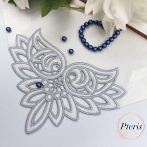 Könnte beinhalten: Ein weiß-graues, gesticktes Blumendesign auf weißem Hintergrund. Das Design ist ein Eckstück mit komplizierten Details. Ein blaues Perlenarmband und verstreute blaue Perlen sind ebenfalls vorhanden.