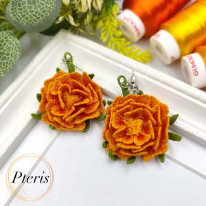 Peut inclure: Paire de boucles d'oreilles artisanales en forme de fleur orange avec des détails verts. Les boucles d'oreilles présentent un motif floral texturé et détaillé. Le mot "Pteris" est dans un cercle sur la surface blanche.