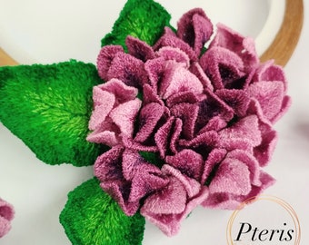 May include: A three-dimensional embroidered hydrangea flower featuring layered purple and pink petals with vibrant green leaves. The embroidery displays dense stitching and texture. The word Pteris is printed in black within a gold circle.