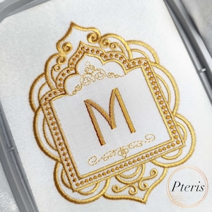 May include: White fabric with an embroidered gold design. The design features a square frame with the letter "M" inside, surrounded by an ornate, gold filigree border. The design is likely for a monogram or personalized item.
