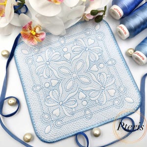 Puede incluir: Un posavasos bordado cuadrado, azul claro y blanco, con un diseño floral y de corazón. El posavasos tiene un borde azul claro y está rodeado de cinta azul, perlas y carretes de hilo azul. Las orquídeas blancas están en el fondo.