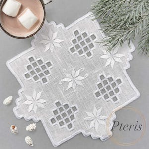 Puede incluir: Doily de crochet blanco con un diseño de copo de nieve. El doily está sobre una superficie gris con elementos decorativos blancos.