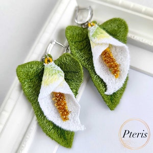 May include: A pair of embroidered calla lily earrings featuring green leaves, white petals, and yellow beaded spadices. The earrings are attached to silver-toned metal lever-back hooks. The word Pteris is printed in the bottom right corner.