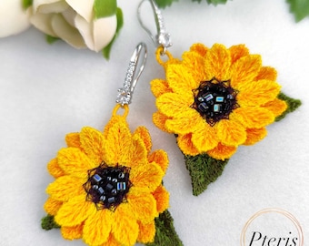 Puede incluir: Un par de pendientes de girasol con pétalos bordados en amarillo, centros oscuros con cuentas negras e iridiscentes, y hojas bordadas en verde. Los pendientes cuelgan de ganchos de metal plateado con pequeñas piedras claras.