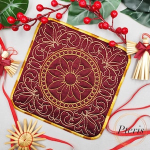 May include: A square, burgundy-colored quilted coaster with a gold floral design. The coaster has a gold border and a central flower motif. The word "Pteris" is visible in the bottom right corner. Decorative Christmas ornaments and red berries are in the background.