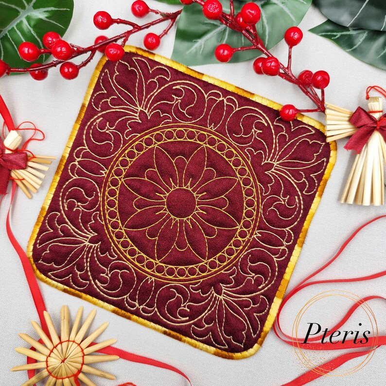 May include: A square, burgundy-colored quilted coaster with a gold floral design. The coaster has a gold border and a central flower motif. The word "Pteris" is visible in the bottom right corner. Decorative Christmas ornaments and red berries are in the background.