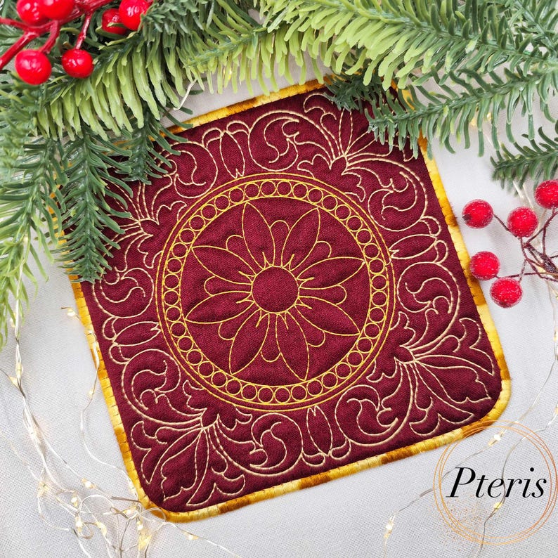 May include: A square, burgundy coaster with a gold floral design. The coaster has a gold border and a central flower motif surrounded by a circular pattern. The design is embroidered. The coaster is a decorative item.