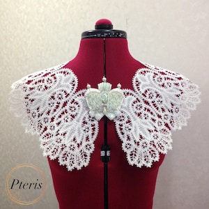 Peut inclure: Un col en dentelle blanche avec un motif floral délicat, présenté sur un mannequin de robe rouge. Une broche décorative avec des pierres irisées est épinglée au col.