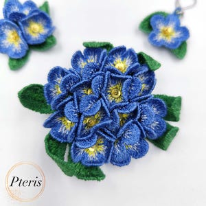 Könnte beinhalten: Eine blaue und grüne gestickte Blumenbrosche mit gelben Zentren. Die Blume besteht aus vielen kleinen Blütenblättern und ist von grünen Blättern umgeben.