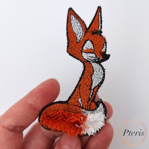 Może przedstawiać: Naszywka z haftowanym pomarańczowym i białym lisem z puszystym ogonem. Naszywka leży na dłoni.