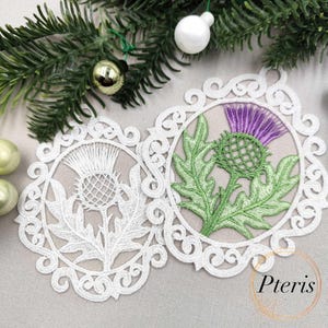 Puede incluir: Dos adornos redondos de encaje blanco con un dise&ntilde;o de cardo. Uno es completamente blanco, el otro presenta un cardo verde y morado. Los adornos est&aacute;n rodeados por un borde decorativo y sobre un fondo neutro.
