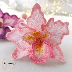 Puede incluir: Una flor de orqu&iacute;dea bordada de color rosa con un centro naranja brillante. La flor est&aacute; hecha de tela y tiene un dise&ntilde;o delicado y en capas.