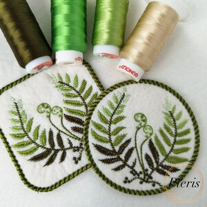 May include: Two embroidered felt coasters with a fern design. The coasters are green and brown and have a green border. The coasters are on a white surface with spools of green, brown, and beige embroidery floss.