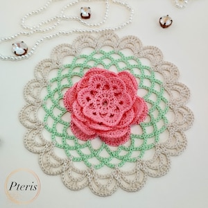 Könnte beinhalten: Eine gehäkelte Tischdecke mit einem zarten Blumenmuster in Beige-, Grün- und Rosatönen. Die Tischdecke hat eine große, mehrschichtige Blume in der Mitte.