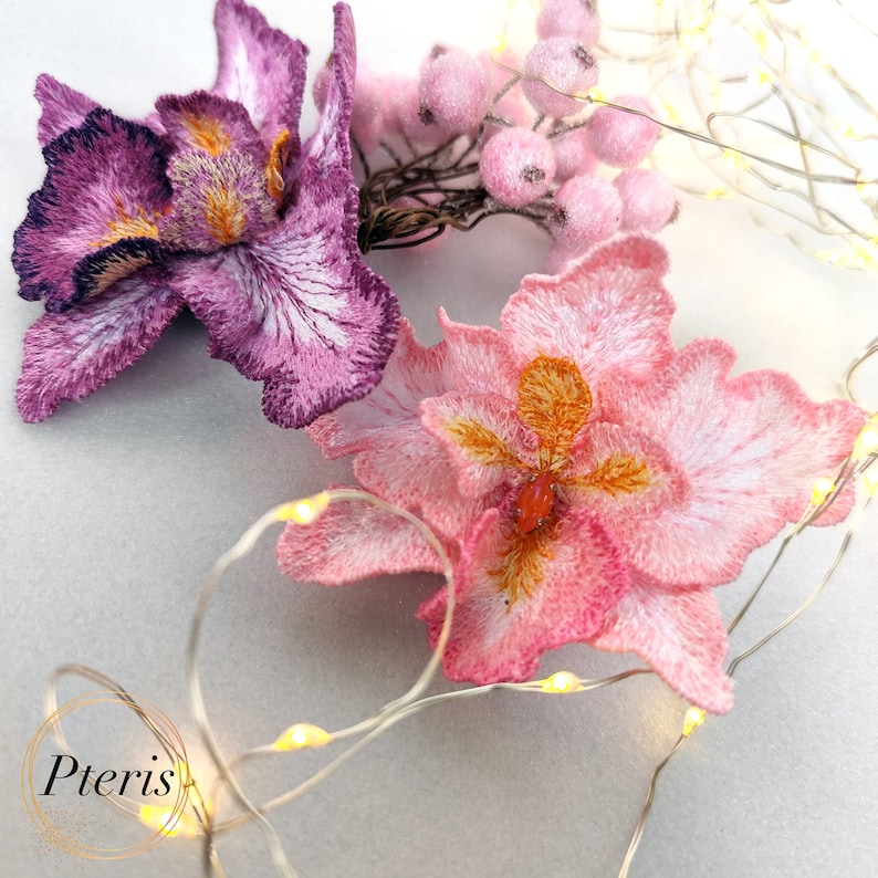Puede incluir: Tres flores bordadas, una morada, una rosa y una rosa p&aacute;lida con un centro amarillo, est&aacute;n dispuestas sobre un fondo blanco con una cadena de luces. El texto "Pteris" es visible en la esquina inferior izquierda.