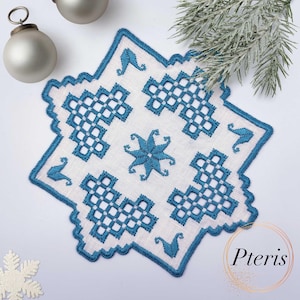 Puede incluir: Un mantel de lino blanco con un diseño de copo de nieve bordado en azul. El mantel tiene un borde festoneado y es perfecto para agregar un toque de elegancia a cualquier mesa.