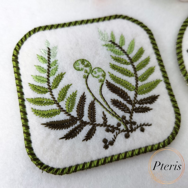 May include: A square green and brown embroidered fern design on a white fabric coaster with a green stitched border.