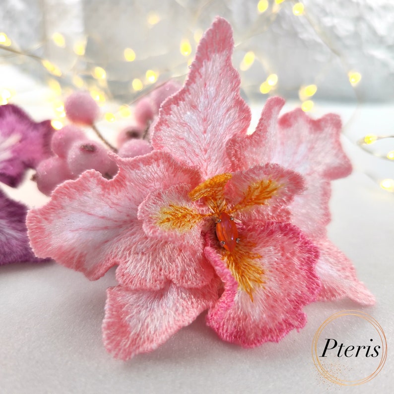 Puede incluir: Una flor de orqu&iacute;dea bordada en rosa y blanco con un centro amarillo. La flor est&aacute; hecha de una tela suave y texturizada.