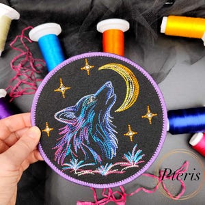 May include: A circular black fabric patch featuring an embroidered design of a wolf howling at a yellow crescent moon. The wolf is stitched in shades of blue, purple, and pink. Four yellow stars and small floral elements surround the wolf.