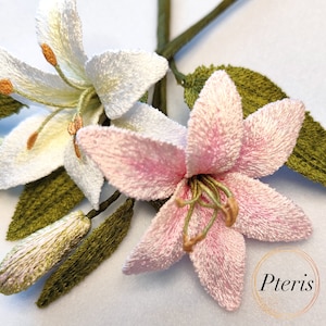 Op de afbeelding: Close-up van twee geborduurde lelies, een witte en een roze, met groene bladeren en een knop. De roze lelie heeft een gedetailleerd centrum met groene meeldraden. De afbeelding is gesigneerd met "Pteris".