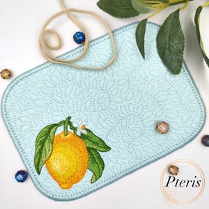 Puede incluir: Un mantel individual acolchado azul claro con un bordado de limón. El limón es amarillo con hojas verdes y una flor blanca. El mantel individual tiene un borde azul claro y un patrón decorativo.