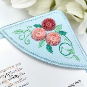 以下が含まれることがあります： 淡い青色の刺繍入りブックマーク。3つのピンクと1つの赤い花、緑の葉、装飾的なつるのデザインです。ブックマークは淡い青色の縁取りで、「Pteris」の文字が入っています。