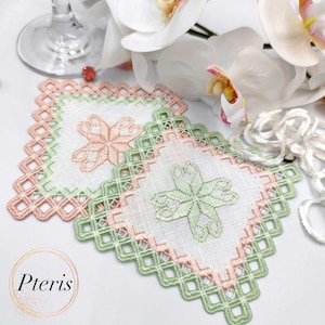 Peut inclure: Deux dessous de verre carrés en lin blanc avec un motif floral. Chaque dessous de verre a une bordure en forme de losange dans des tons alternés de vert clair et de pêche. Les dessous de verre sont placés sur une surface blanche, avec un verre à vin et des fleurs en arrière-plan. La marque "Pteris" est dans le coin inférieur gauche.